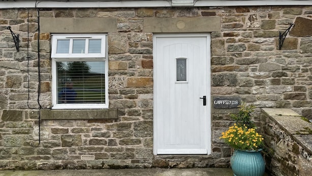 A stone building with a white door and window, a sign reading "CAWFIELD," and a potted plant with yellow flowers. - Milecastle Inn on Hadrian's Wall Near Haltwhistle (Haltwhistle)
