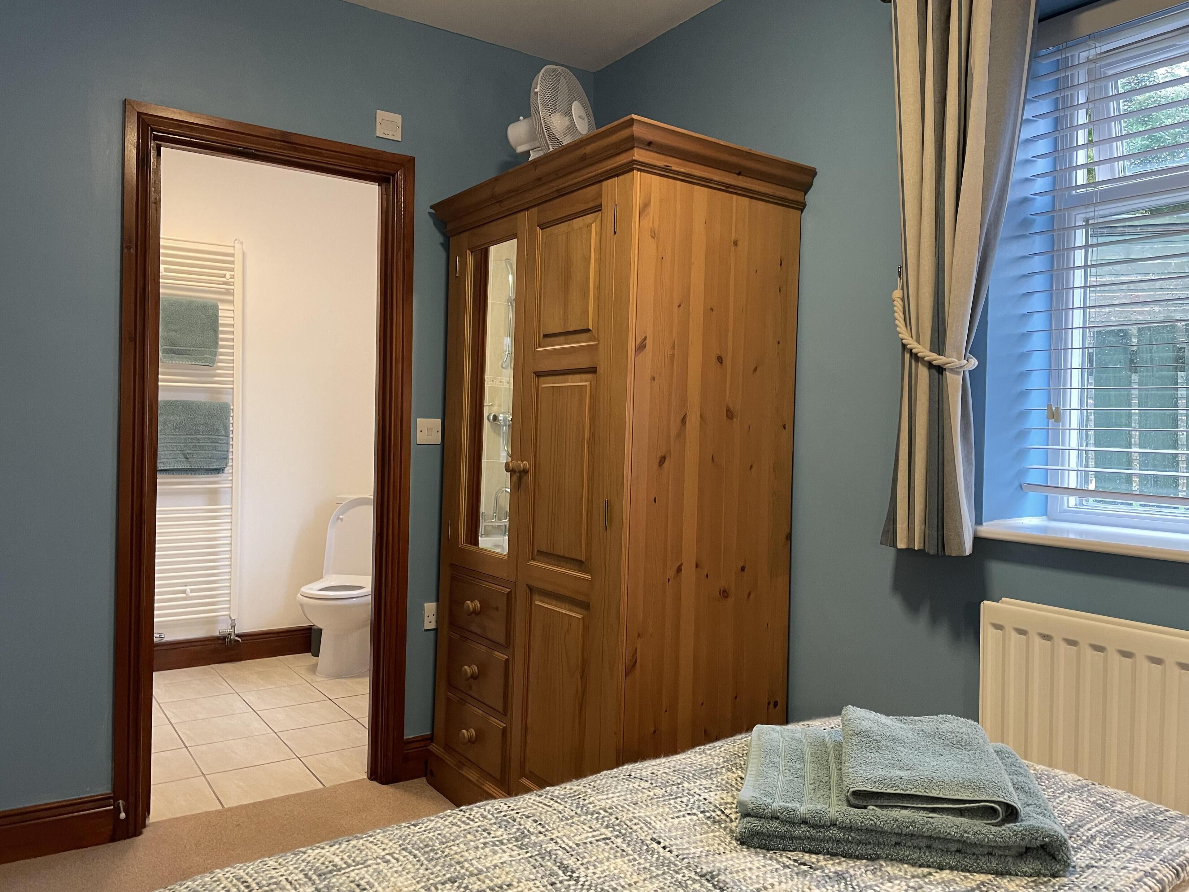 Een slaapkamer met een houten garderobe, een bed met een gebreid plaid, een raam met zonwering, en uitzicht op de badkamer.