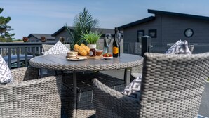 Een ronde buitentafel met rieten stoelen, een bloempot, een fles champagne en een bord met eten en drinken.