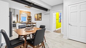 Uma zona de refeições moderna com mesa de madeira e cadeiras pretas, cozinha ao fundo e parede amarelo vibrante.