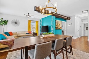 A modern living room with a large wooden dining table, comfortable seating, a flat-screen TV, and a unique wall design.