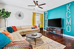 A living room with a sectional sofa, a round coffee table, a television, and a wall-mounted clock. - Belvedere Flats - 2 units + Walkable to Westport! (Kansas City)
