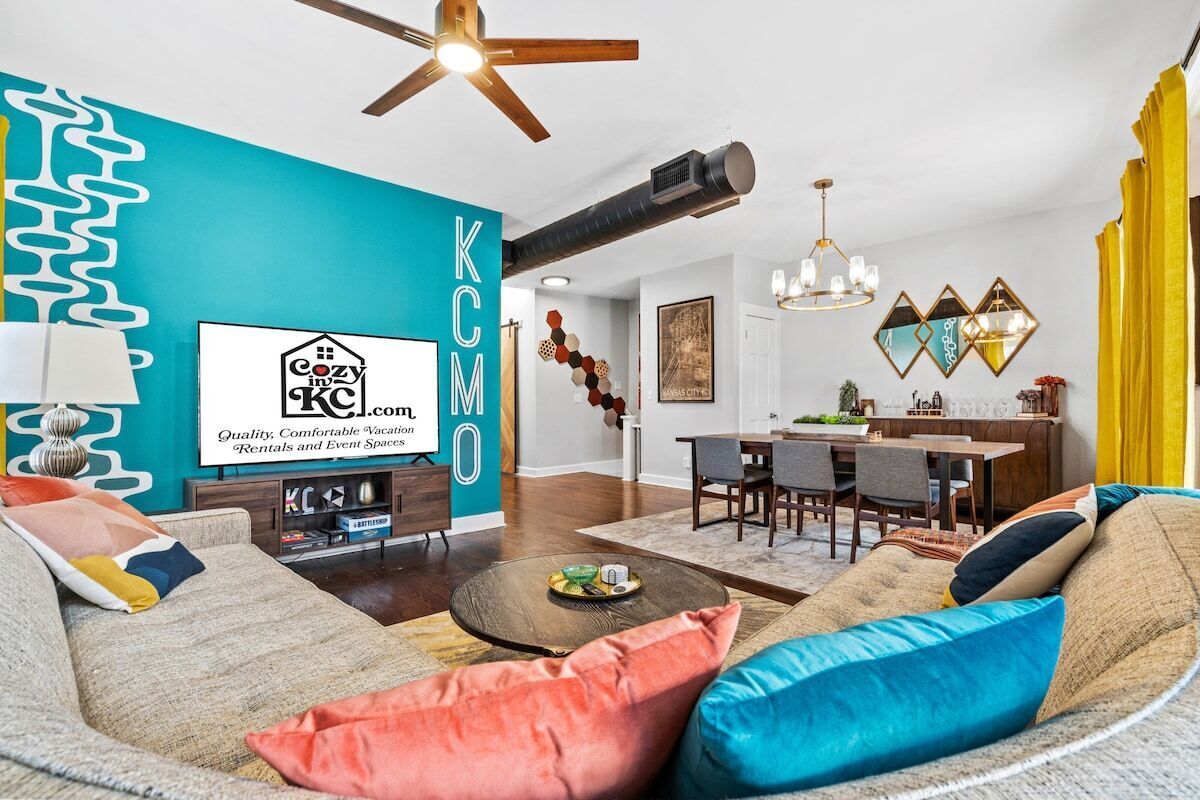 A modern living room with a teal accent wall, a ceiling fan, a dining area, and a kitchen visible in the background.