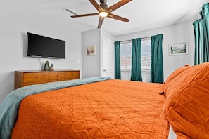 Une chambre à coucher comprenant un lit, un ventilateur de plafond, une télévision, une commode et une fenêtre avec des rideaux.