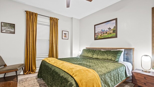 A bedroom with a bed, a nightstand, a chair, a window with curtains, and a painting on the wall.