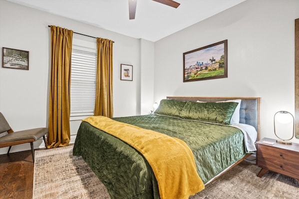 A bedroom with a bed, a nightstand, a chair, a window with curtains, and a painting on the wall.