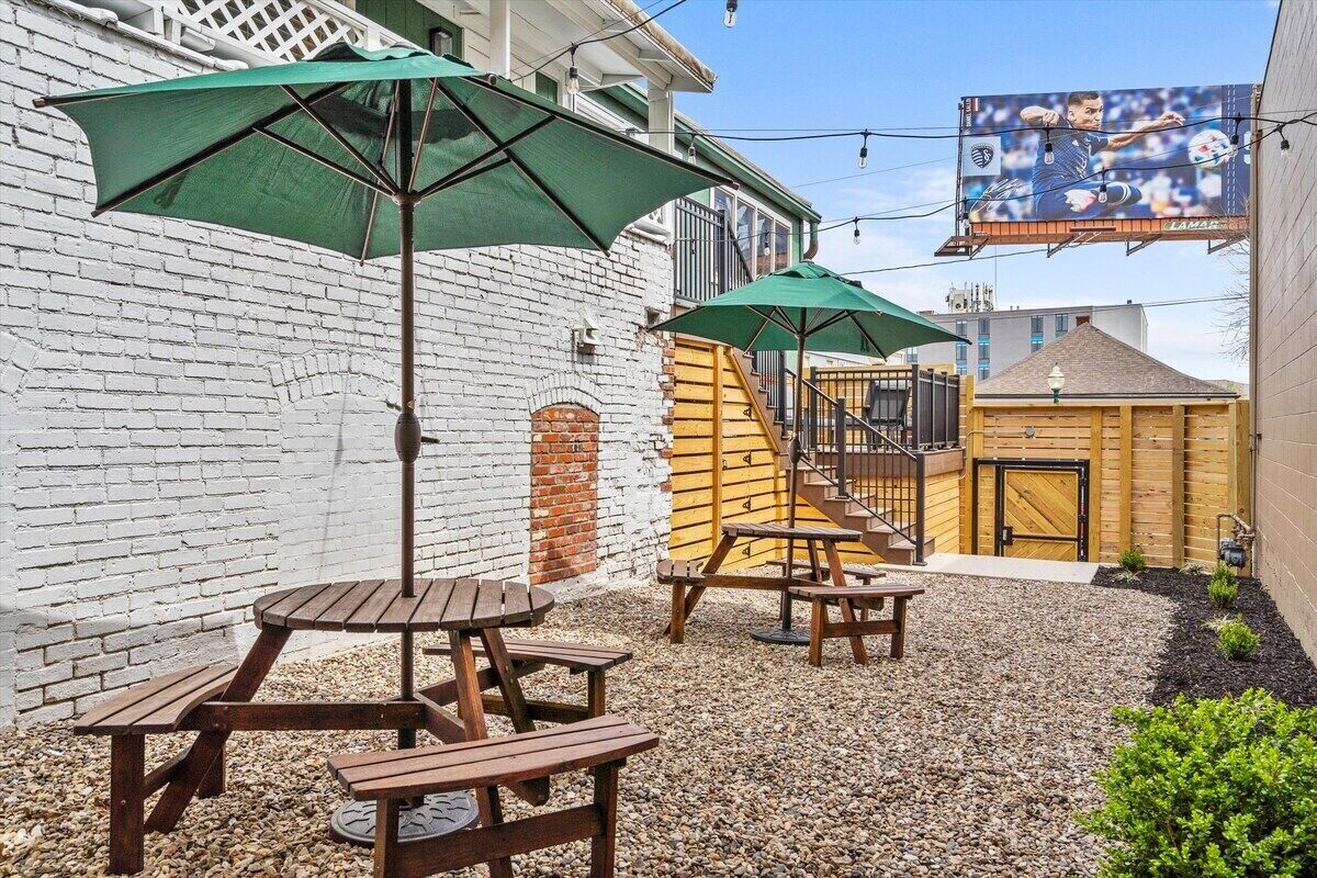 A patio area with wooden tables and green umbrellas, a brick wall, and a wooden building in the background.