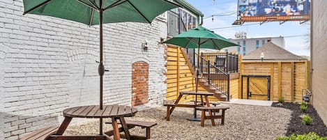 A patio area with wooden tables and green umbrellas, a brick wall, and a wooden building in the background.