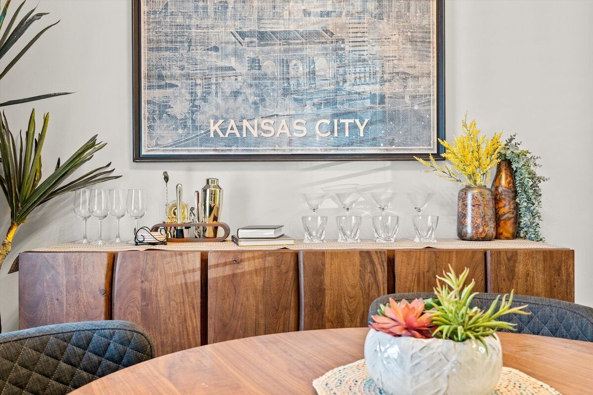 A dining area with a round table, chairs, a wooden cabinet with glassware, and a framed cityscape artwork.