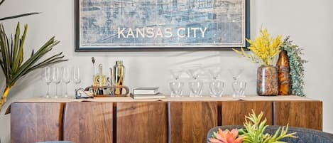 A dining area with a round table, chairs, a wooden cabinet with glassware, and a framed cityscape artwork.