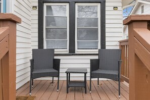 A wooden deck with two wicker chairs and a small table, a window with blinds, and a white exterior wall. - Waldo Craftsman - Fenced yard + Walkable to dining (Kansas City)