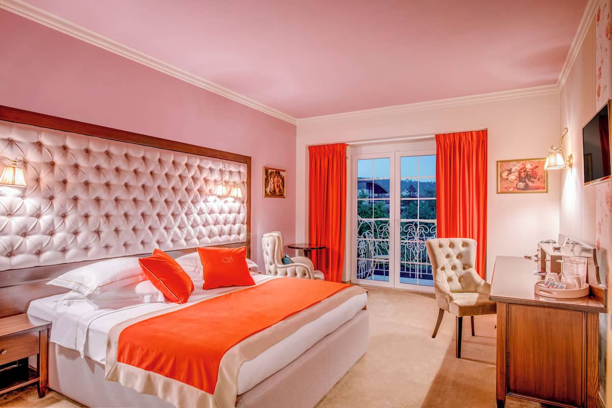 A bedroom with a tufted headboard, a bed with white and orange bedding, a desk with a coffee maker, and a view of a balcony with red curtains.
