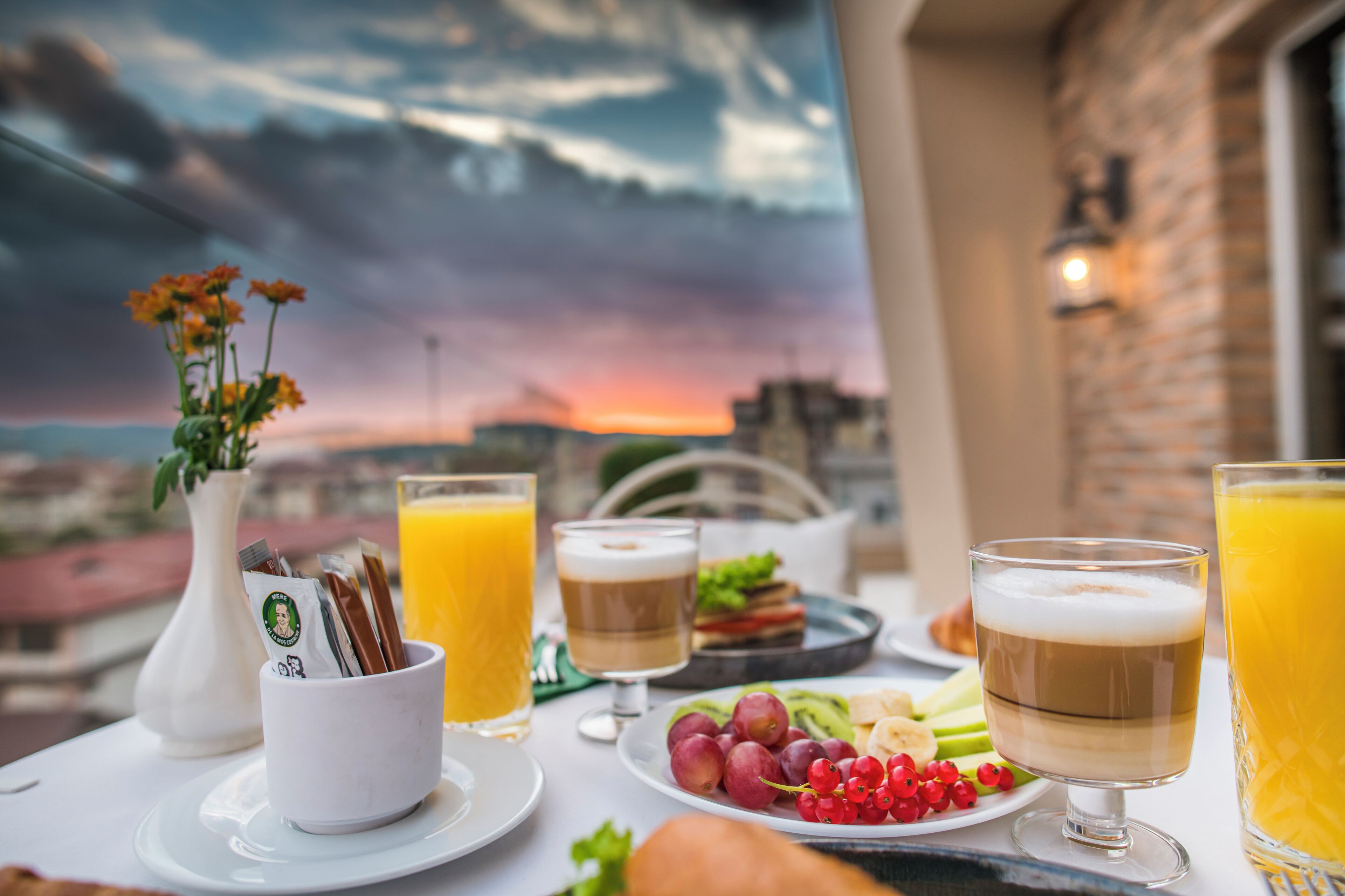 Une table de petit-déjeuner avec vue sur la ville, agrémentée d’un vase de fleurs, d’une assiette de fruits et de verres de jus d’orange et de café.