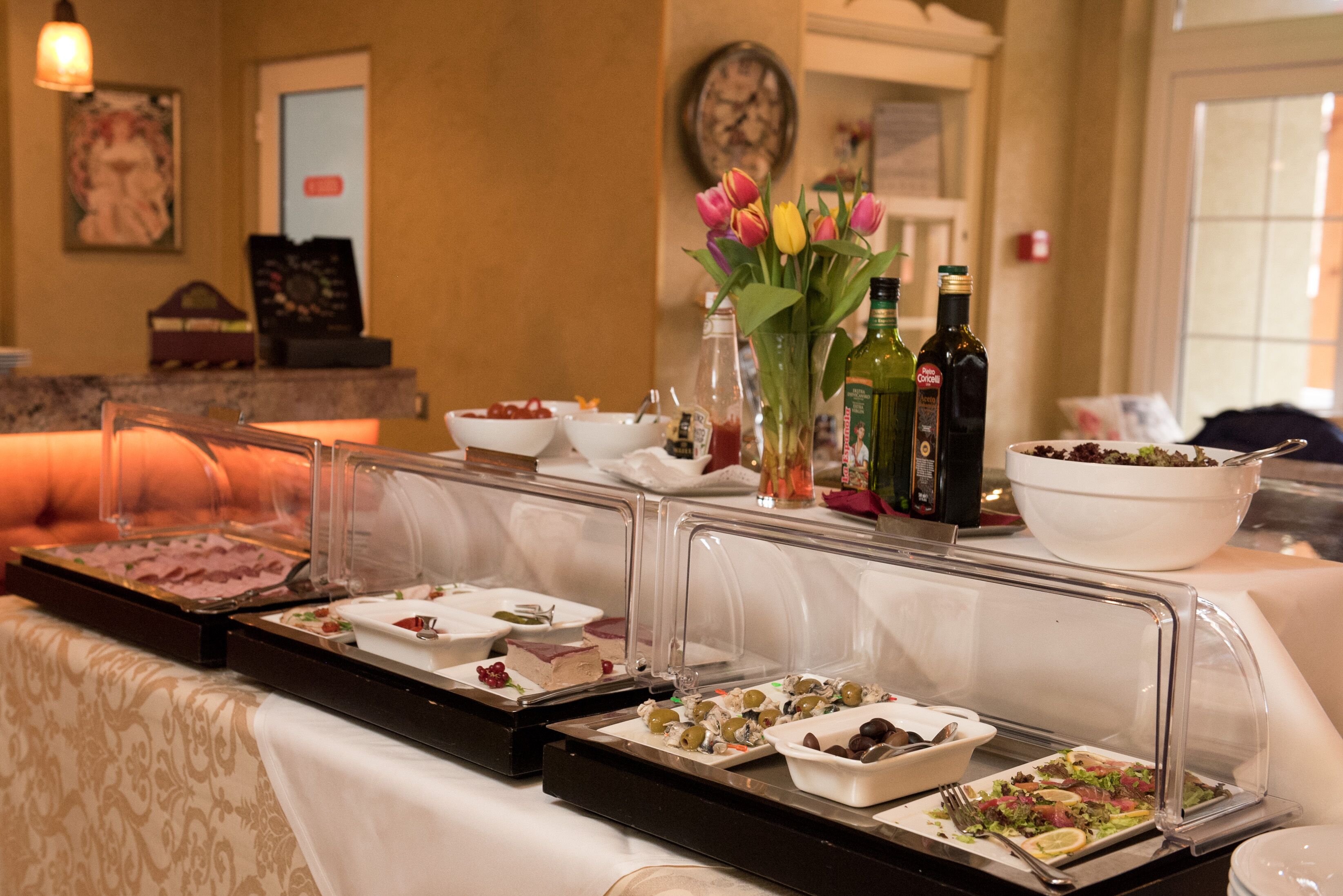 A buffet setup with various food items and drinks, a vase with flowers, and a clock on the wall.