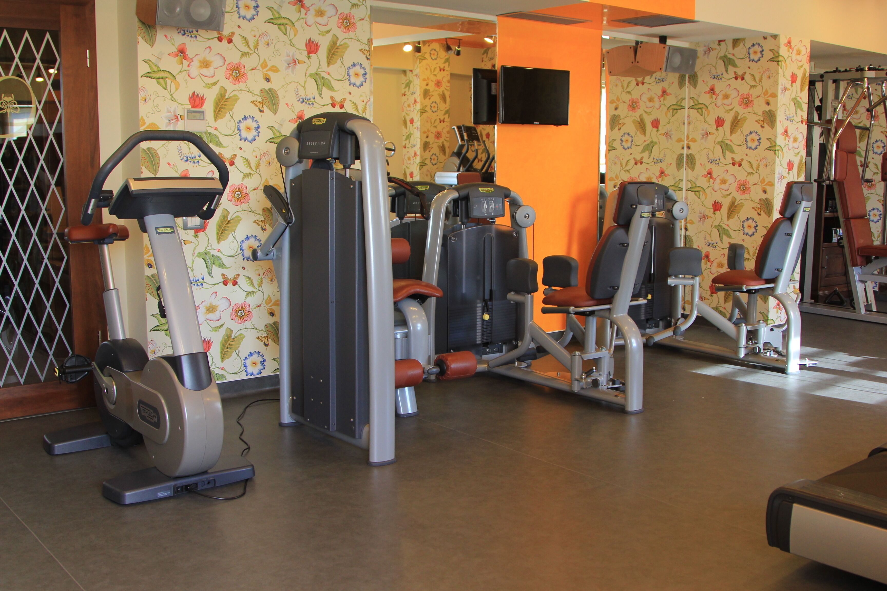 Un gimnasio con diversas máquinas de ejercicio, un televisor instalado en la pared y un diseño de papel tapiz con motivos florales.