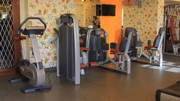 Un gimnasio con diversas máquinas de ejercicio, un televisor instalado en la pared y un diseño de papel tapiz con motivos florales.