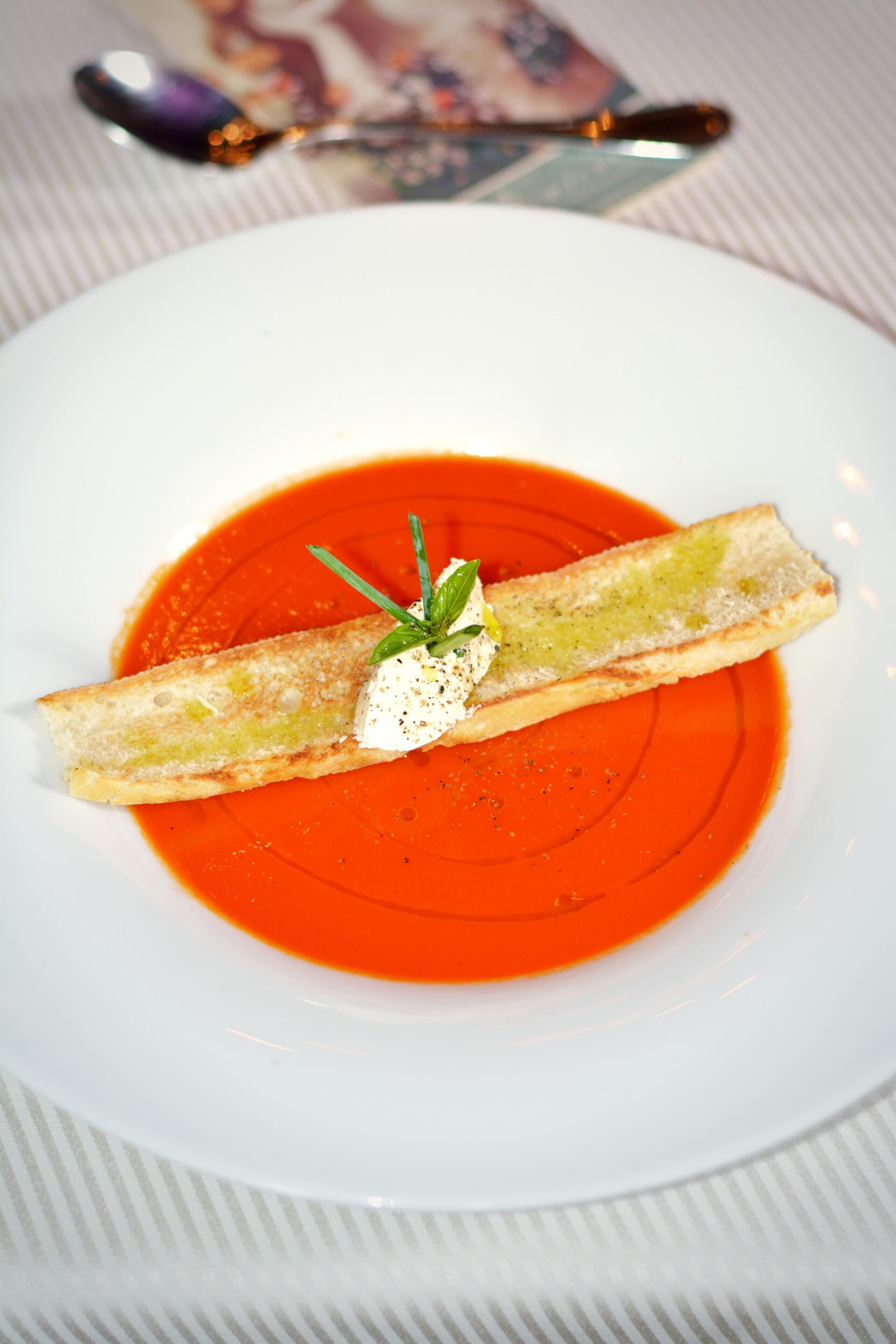 A white plate with a slice of bread topped with a dollop of cream and a sprig of herb, placed on a bed of red sauce.