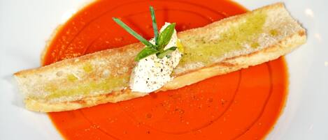 A white plate with a slice of bread topped with a dollop of cream and a sprig of herb, placed on a bed of red sauce.