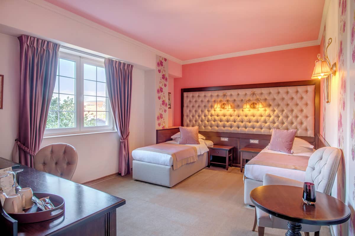 A hotel room with a large bed, a smaller bed, a desk with a chair, a table, and a window with curtains.