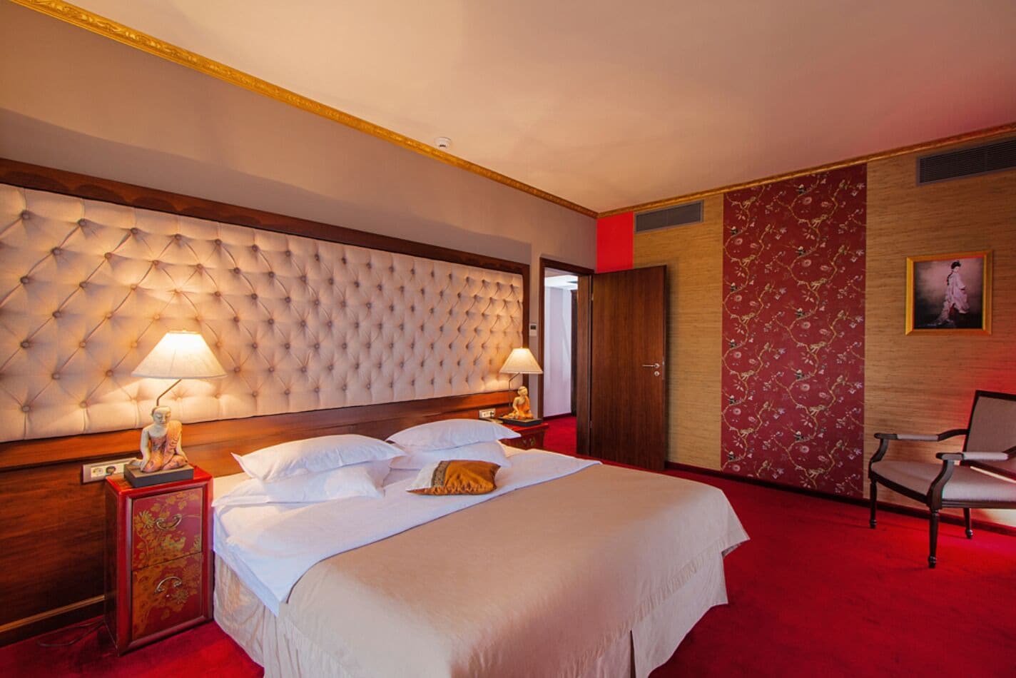 A hotel room with a tufted headboard, a bed with white linens, two bedside lamps, a wooden nightstand, a red carpet, and a chair.