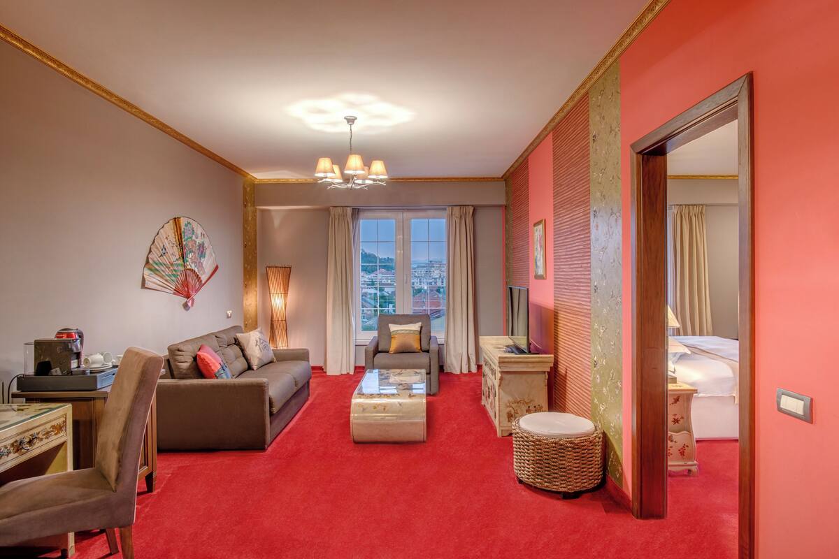 A hotel room with a red carpet, a sofa, a chair, a coffee table, a lamp, a mirror, and a view of the city through the window.