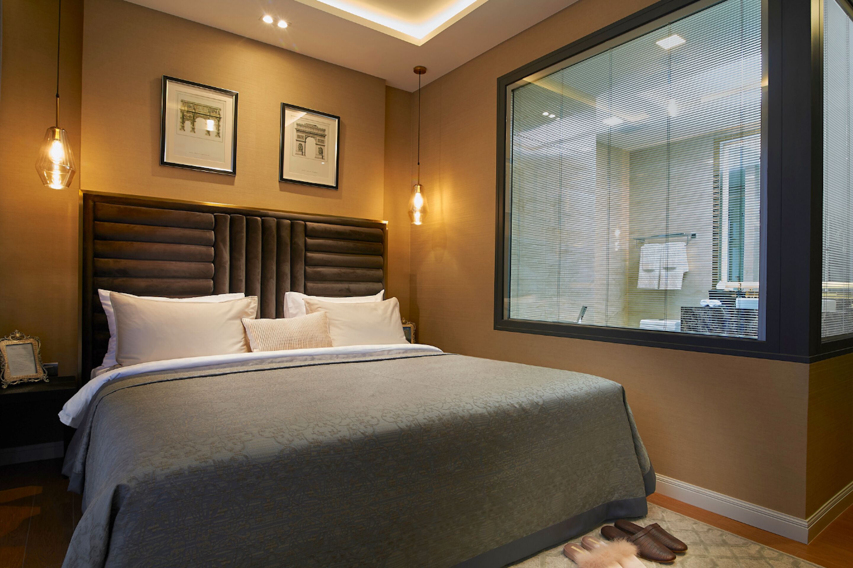 Une chambre moderne avec un grand lit, une tête de lit à lattes verticales, deux tableaux encadrés au mur et une grande fenêtre avec des stores.
