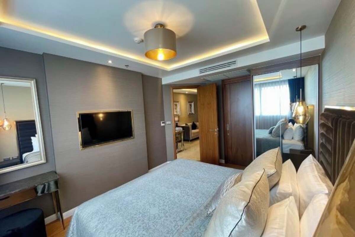 A modern hotel room with a large bed, a flat-screen TV mounted on the wall, and a mirror above the bed.