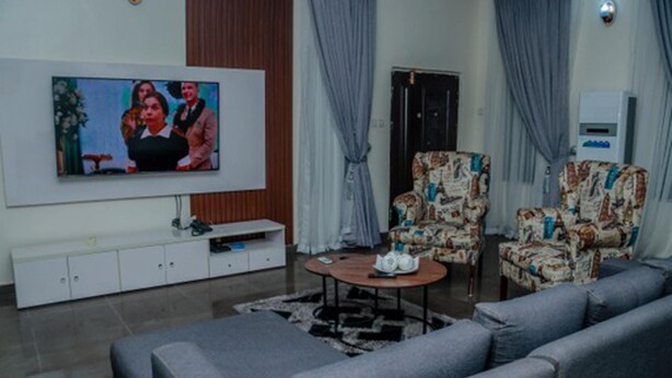 Uma sala de estar com televisão de tela plana, sofás cinzentos, poltronas estampadas, uma mesa de centro e um rack branco.