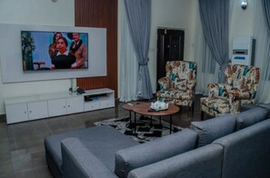 A living room with a flat-screen TV, grey sofas, patterned armchairs, a coffee table, and a white entertainment unit. - Stunning 3-bed House in Sapele (Sapele)