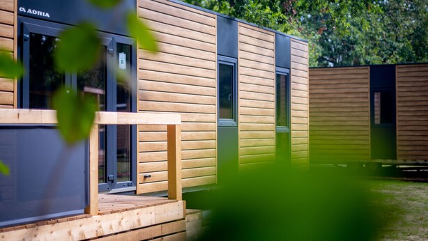 Een modern houten gebouw met een glazen deur en een houten terras, met een bord waarop 'ADRIA' staat.