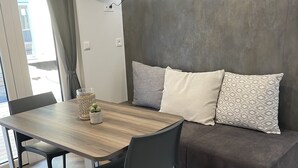 Een moderne woonruimte met een houten eettafel, twee stoelen, een bank met gekleurde kussens en een wandgemonteerde flatscreen-tv.