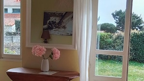 Ett rum med ett träbord, en vas med blommor, en tavla på väggen och en utsikt över grönska genom en dörr.