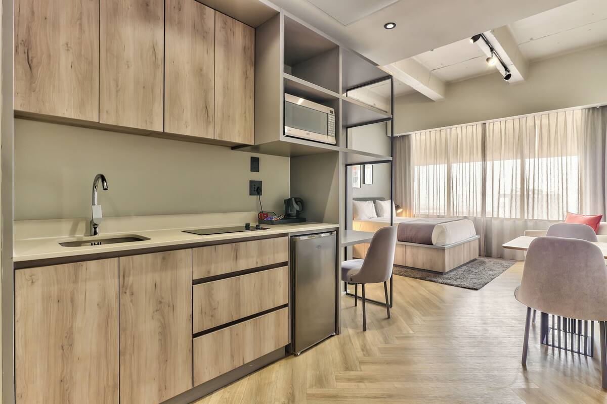 A modern kitchen with wooden cabinets, a built-in microwave, and a refrigerator. There is a dining area with a sofa and chairs, and a large window with curtains.