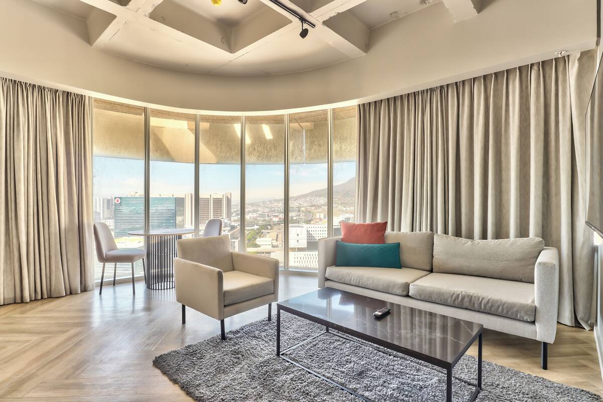 A modern living room with a sofa, armchair, and coffee table, large windows with curtains, and a city view.