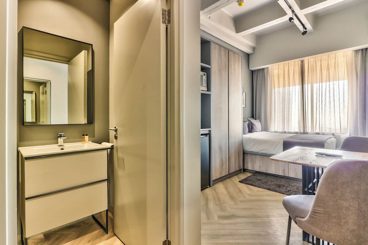 A modern hotel room with a bathroom featuring a sink and mirror, a bed with a wooden headboard, a small table with a chair, and a large window with curtains.