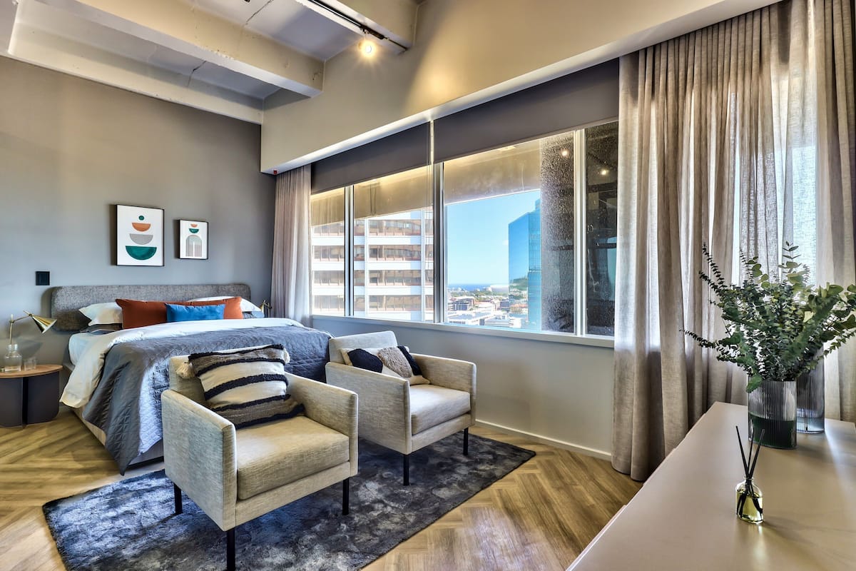 A modern bedroom with a large bed, two armchairs, a nightstand, a large window with curtains, and a city view.