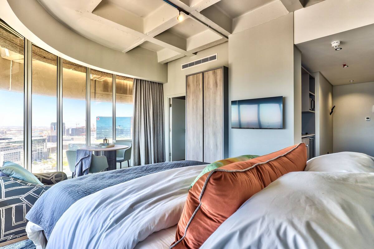 A modern hotel room with a curved ceiling, large windows offering a city view, a bed with multiple pillows, a small table, and a chair.