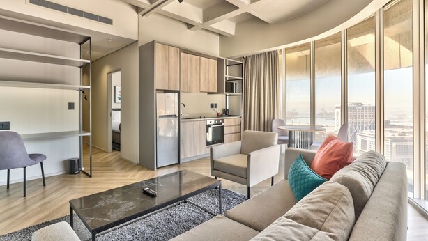 Un appartement moderne comprenant un salon, une cuisine et un coin repas. Il dispose d’une grande fenêtre offrant une vue sur la ville, d’un canapé avec des coussins et d’une table basse.
