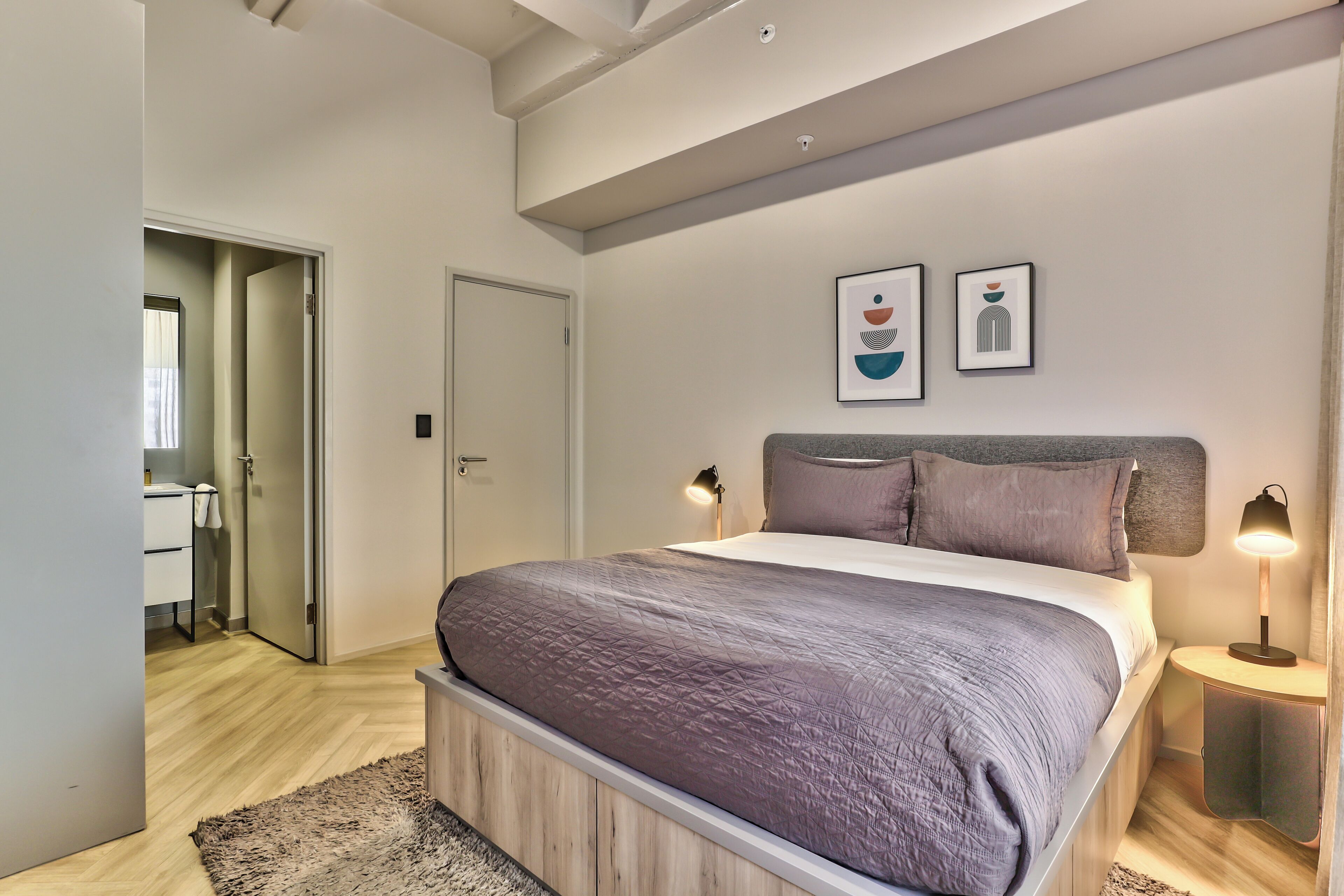 A modern bedroom with a large bed, a bedside table with a lamp, and a door leading to a bathroom.