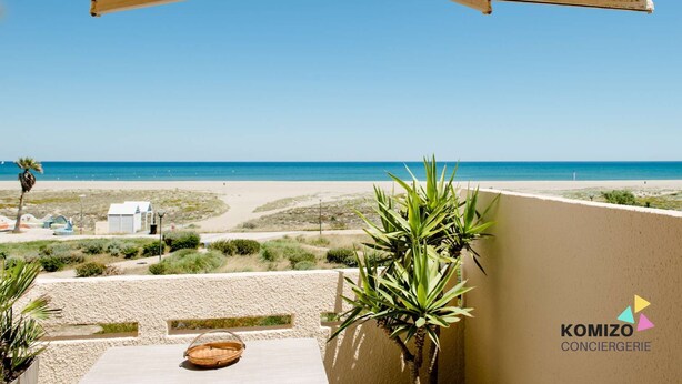 Een balkon met uitzicht op het strand, een palmboom en een klein tafeltje met een mand.