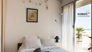 Hotelkamer met een bed, kussens, een bedlampje, een ingelijst kunstwerk aan de muur en uitzicht op het strand door het raam.