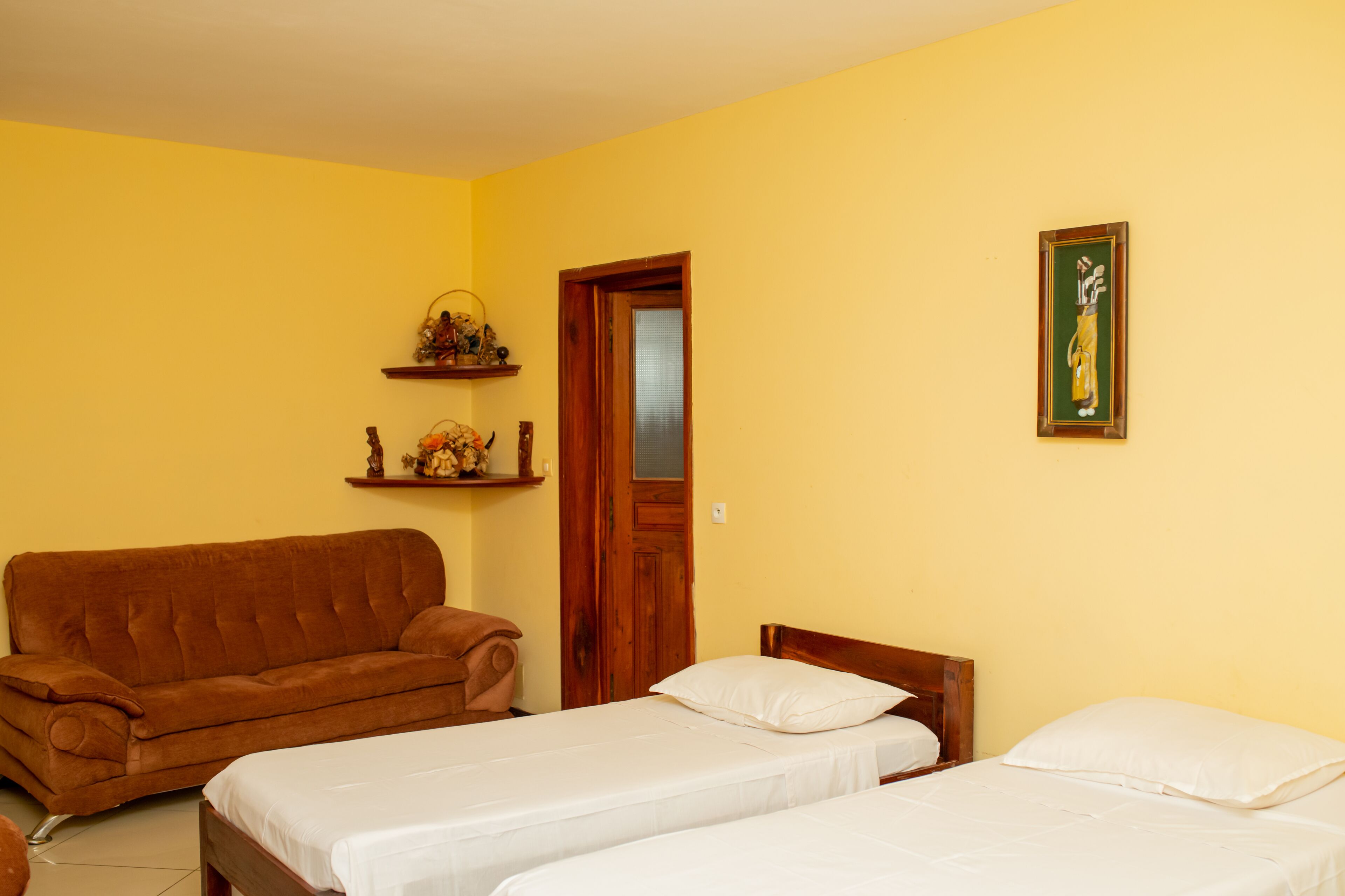 A room with two single beds, a brown sofa, a wooden door, and a wall-mounted shelf with decorative items.
