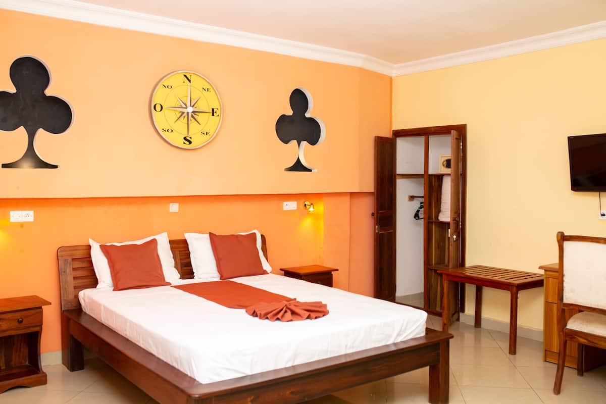 A hotel room with a large bed, wooden headboard, and a wall clock. There is a wooden chair and a small table beside the bed.
