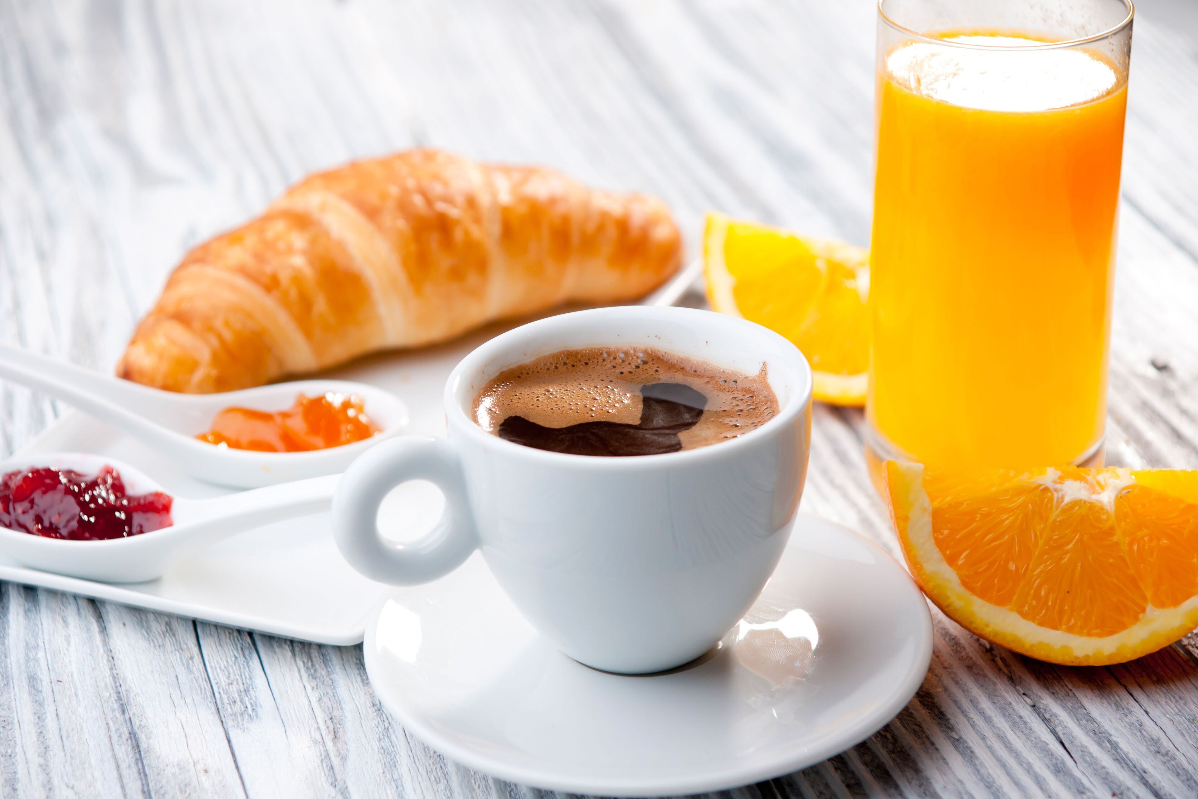 Morgenmadsanretning med croissant, kaffekop, et glas appelsinjuice og en appelsinskive på en hvid tallerken.