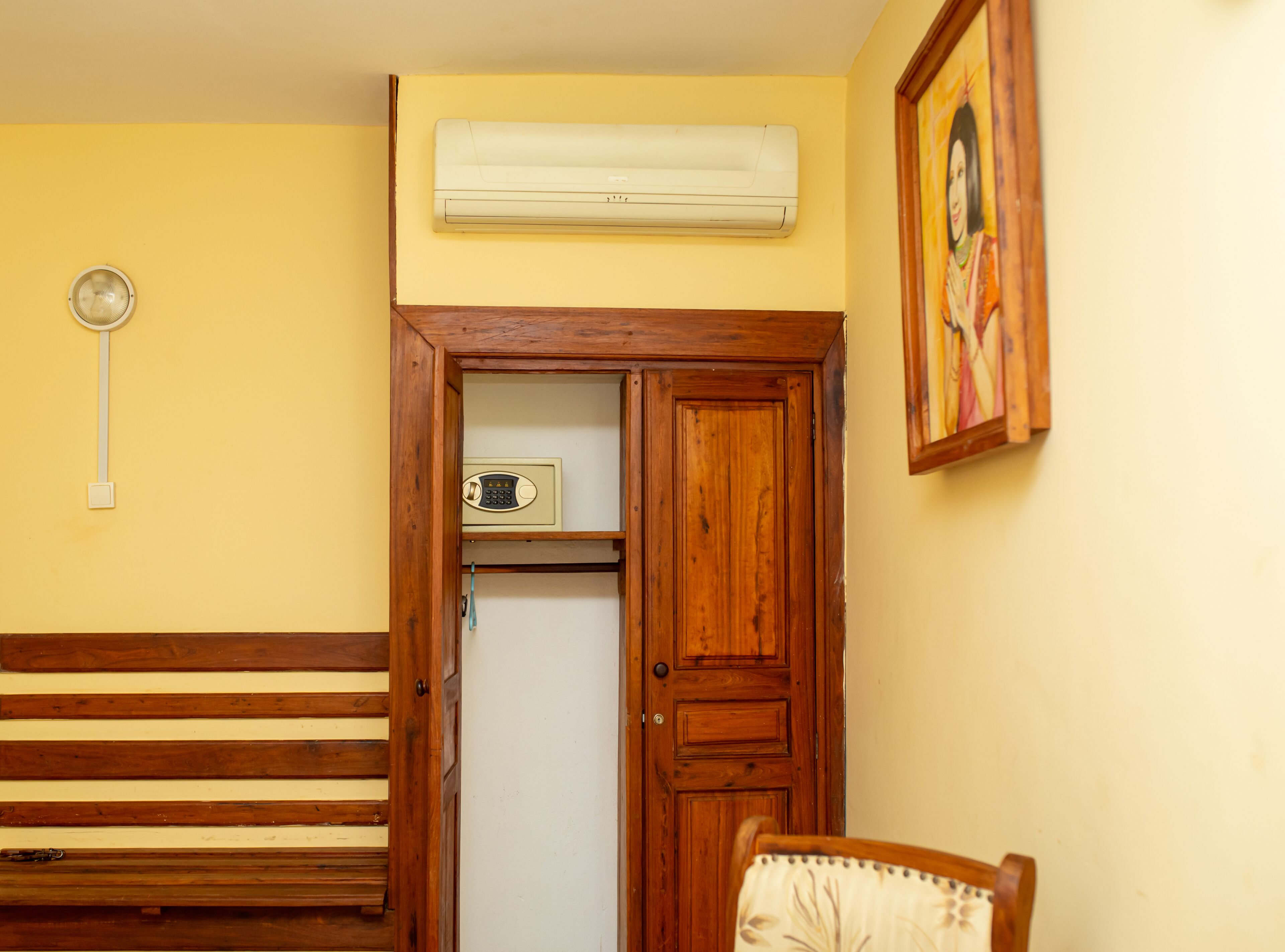 A room with a wooden bench, a white air conditioner mounted on the wall, a small shelf with a radio, and a framed picture on the wall.