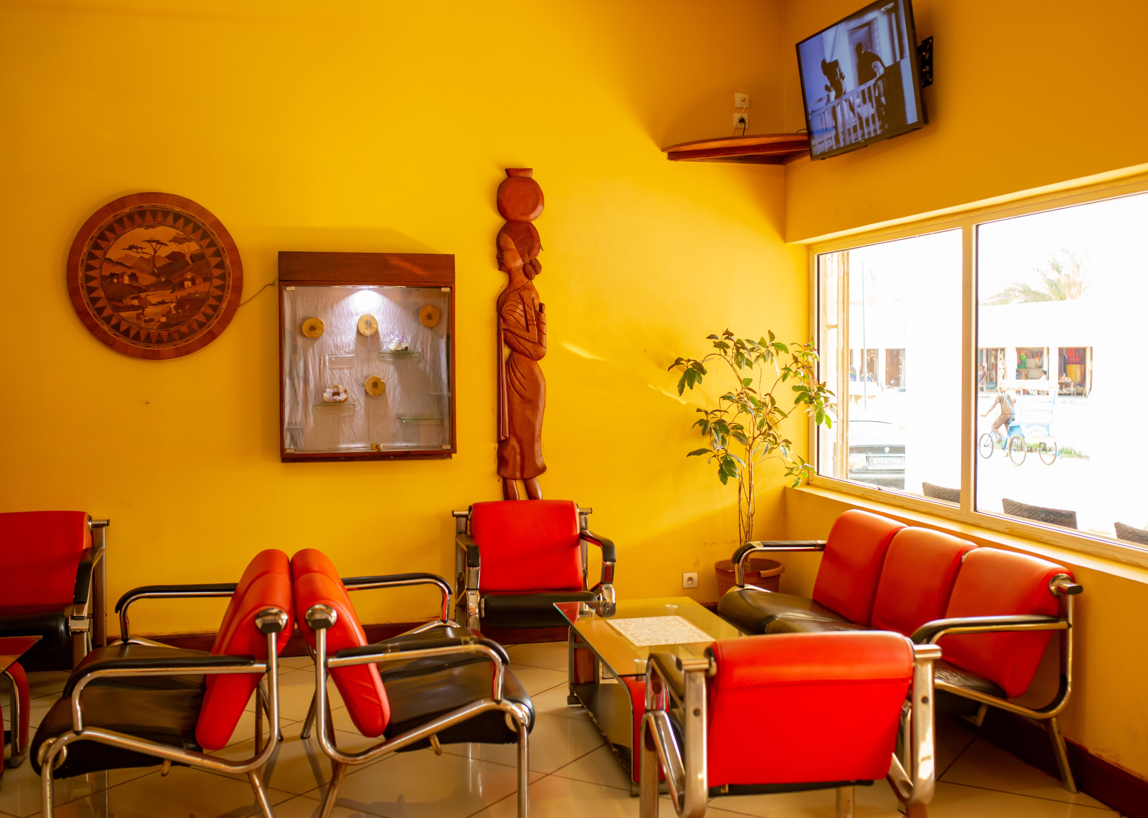 A waiting area with red chairs, a wooden statue, a wall-mounted TV, and a window with a view of the street.