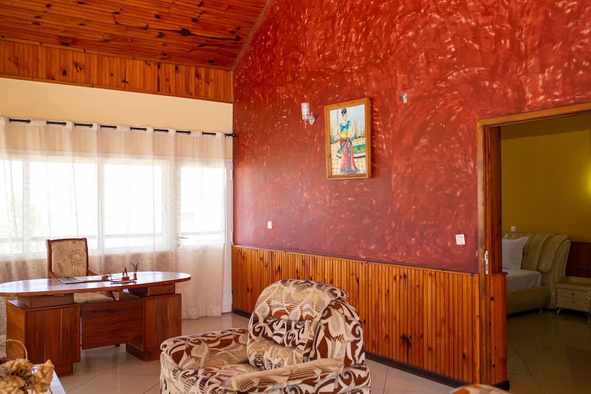 A room with a wooden ceiling, a desk with a chair, a patterned armchair, and a painting on the wall.