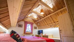 A cozy attic bedroom with a sloped ceiling, a bed with checkered bedding, a window with red curtains, and a television on a wooden dresser.