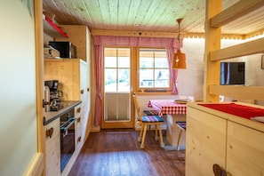 Un acogedor interior de cabaña con paredes de madera, una zona de cocina, una mesa de comedor con mantel a cuadros y una ventana con cortina de cuadros rojos y blancos.