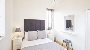 A modern bedroom with a large bed, a nightstand with a lamp, a small stool, a window with blinds, and a mirror on the wall. - Host & Stay | Boutique Balcony (Whitby)